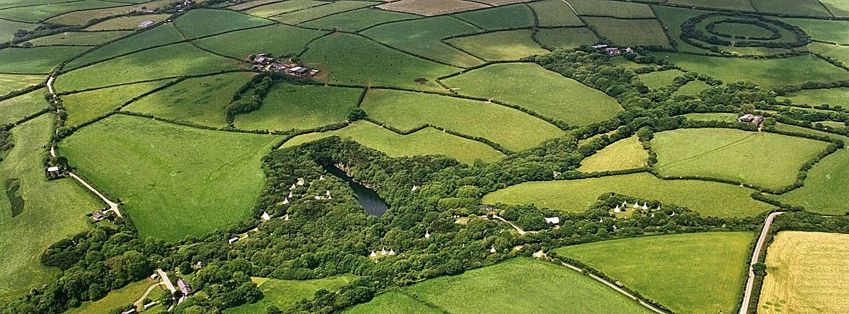 Aerial photograph of tipis tucked into woodland around a dark quarry lake, among green Cornish fields