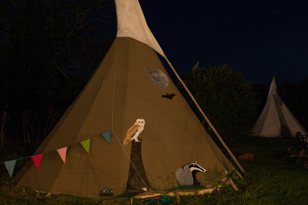 A tipi at night painted with an owl, a moon and a badger